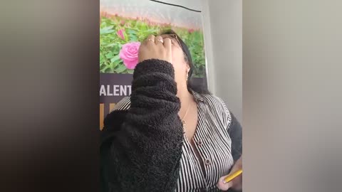 A video shows a person with light skin and short dark hair, wearing a striped shirt, covering their face with both hands, standing in a room with a green plant and pink rose poster in the background.