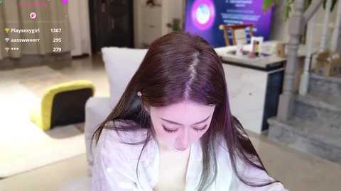 Video of a young Asian woman with long brown hair, wearing a white robe, sitting on a white chair in a modern, well-lit living room with a TV displaying a purple screen.
