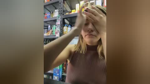 Video of a blonde woman with light skin, wearing a sleeveless pink top, covering her face with her hand, in a messy, cluttered pharmacy with shelves full of products.