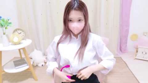 Video of a young Asian girl in a white shirt, kneeling on a wooden floor, wearing a pink mask, surrounded by pastel decor, with a toy clock and stuffed animals.
