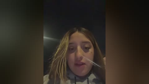 A video of a young woman with long, straight brown hair and pale skin, her mouth covered with a clear surgical mask, set against a dimly lit background.