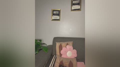 Video of a cozy, dimly-lit room with a grey couch, two framed \"BUSTE\" signs, a plush pink bunny pillow, and a green plant in the corner.