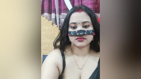 Video of a South Asian woman with medium skin tone, wearing a black mask with floral patterns, red lipstick, and a black spaghetti strap top. She has a gold necklace and sits against a textured yellow wall and red curtains.