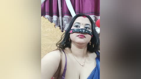 Video of a woman with medium skin tone, wearing a blue dress, and a black-and-white patterned tape over her eyes, standing in front of a golden and red-patterned curtain.