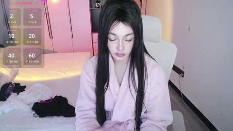 A video of a young Asian woman with long black hair, wearing a light pink robe, sitting on a bed. The background features a white chair, pink wall, and a digital timer display.