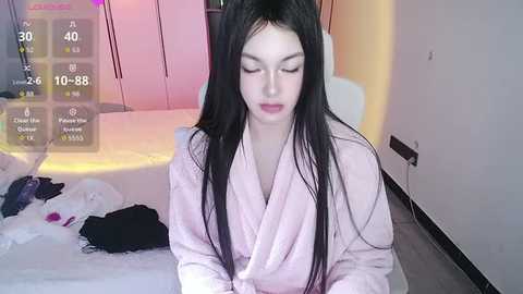A video of a young East Asian woman with long black hair, wearing a light pink robe, sitting on a white bed, surrounded by scattered clothes and a digital fitness screen.