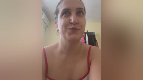 A video of a middle-aged woman with light skin, wearing a red tank top, standing in a dimly lit room with yellow walls and an air conditioner.