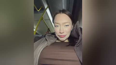 Video of a woman with light skin, straight brown hair, and light makeup, wearing a brown shirt and gray jacket, smiling. Background shows a bus interior with a yellow railing.