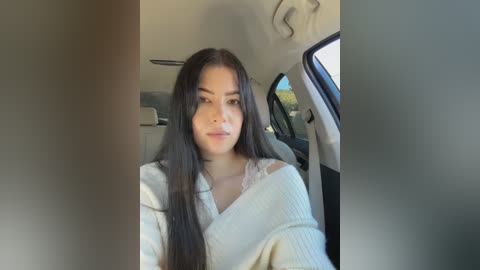 Video of a young woman with long black hair and light skin, wearing a cream-colored sweater, sitting in a car with the door open, showing a kitchen interior in the background.