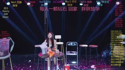 A video of a female singer seated at a small round table, surrounded by colorful stage lights, with a backdrop displaying Chinese text and numbers.