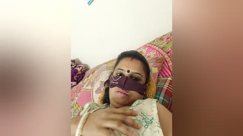 A video of a woman with medium skin tone and dark hair, wearing a purple bindi and a colorful saree, lying on a plaid-patterned pillow in a cozy room.