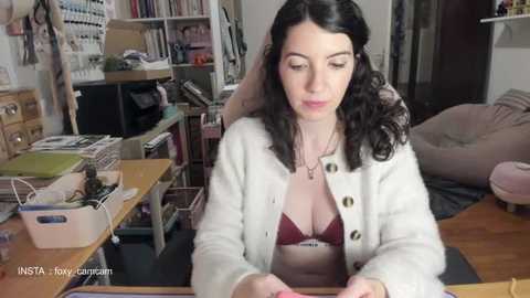 Video of a woman with curly black hair, wearing a white cardigan over a red bra, sitting at a cluttered wooden desk in a messy room with shelves, bins, and a pink beanbag chair in the background.