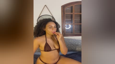 Video of a Black woman with curly hair, wearing a brown bikini, sitting on a bed, biting her finger, in a bedroom with a window and a decorative wall hanging.
