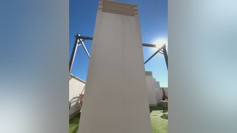Video of a tall, narrow, white, concrete tower with a triangular top, set against a clear blue sky, surrounded by green grass and metal framework.