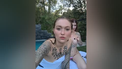 Video of a young woman with intricate black tattoos on her arms and chest, lying on a white towel by a pool, surrounded by lush greenery.