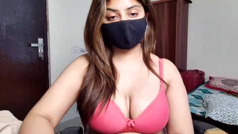 Video of a young woman with long brown hair, wearing a black face mask and pink bra, seated indoors in a dimly lit room with a red couch and patterned blanket.