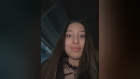 A video of a young woman with long dark hair, light skin, and a slight smile, wearing a black blouse. The background is dimly lit, suggesting an indoor setting.