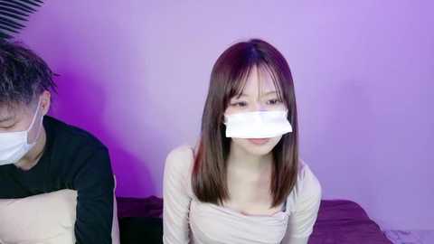 Video of an East Asian woman with straight, shoulder-length brown hair, wearing a white face mask and a beige top, seated on a bed with a man in the background, under purple lighting.