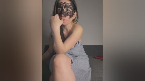 Video of a young woman with light skin and long brown hair, wearing a black lace masquerade mask and gray dress, sitting on a couch in a dimly lit room.