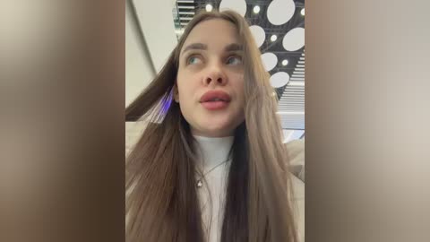 Video of a young woman with long, straight brown hair, styled with a blue headband, wearing a white turtleneck sweater. Background features a black and white polka-dot patterned wall and blinds.