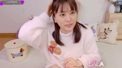 A video of a young Asian woman with straight brown hair, wearing a white, fluffy sweater adorned with teddy bears, sitting at a table in a brightly lit room.