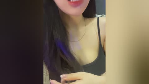 A video of an Asian woman with long black hair, wearing a black tank top, smiling slightly, with a necklace. Her fair skin and pink lipstick are visible.