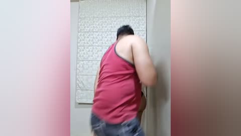 Media: Video of a middle-aged man with a short black beard, wearing a red tank top and blue shorts, pressing against a beige wall with a tiled pattern.