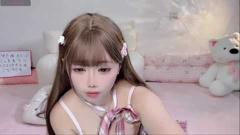 Video of a young Asian woman with long brown hair, wearing a pink and white striped outfit, lying on a bed. The background features a plush teddy bear and a pink quilt.