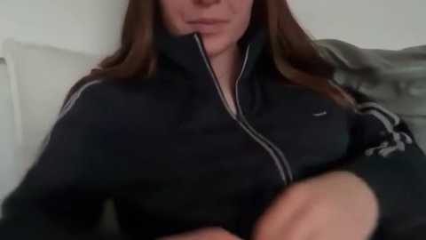 Video of a woman with long brown hair, wearing a black zip-up jacket, sitting on a couch with a neutral-colored background.