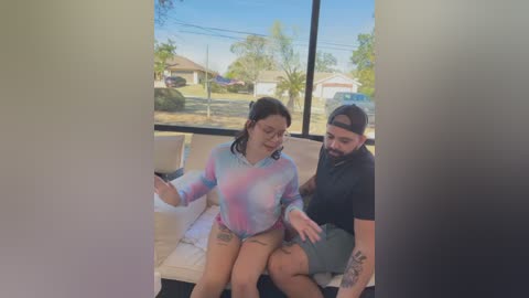 Video of a tattooed woman in a tie-dye top and shorts, sitting on a bench next to a bearded man in a black cap and shirt, outside a window overlooking suburban houses.