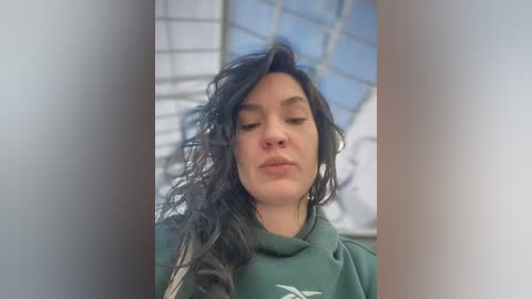 Video of a young woman with wavy black hair, fair skin, and a neutral expression, wearing a green hoodie, in a blurred, glass-paneled indoor setting.