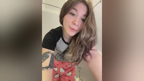 A young woman with long, wavy brown hair, fair skin, and a tattoo on her arm, wearing a black and gray graphic T-shirt with Hello Kitty shorts, posing in a dimly lit bathroom.