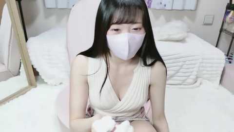 Video of an Asian woman with long black hair, wearing a white face mask, beige ribbed tank top, and pink shorts, sitting on a plush white chair in a cozy bedroom with white bedding and a wooden mirror.