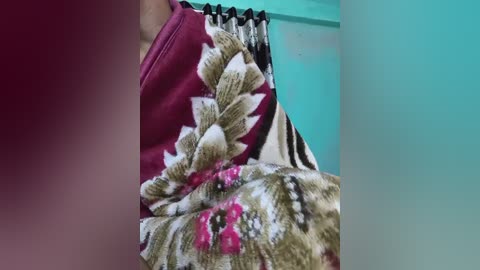 A close-up video of a woman's shoulder, featuring a vibrant, floral-patterned blouse in shades of pink, white, and beige. The background shows a teal wall with a black curtain with white flowers.