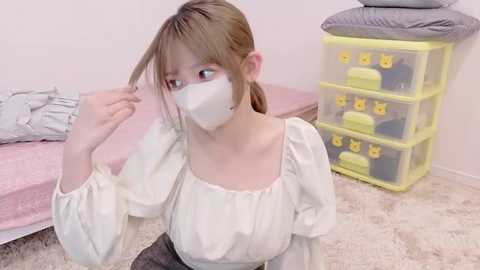 A video of an Asian woman with fair skin and brown hair, wearing a white face mask and white blouse, adjusting her hair in a pink room with a bed, yellow storage unit, and fluffy carpet.