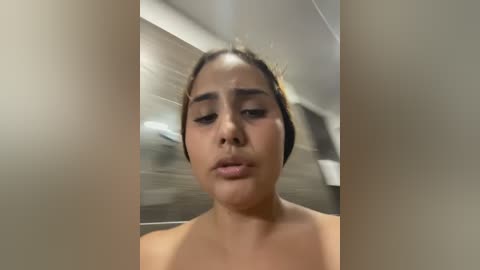 Media: Video of a young woman with medium-brown skin and dark hair, wearing a distressed expression, in a blurry indoor setting with wooden and metallic textures.