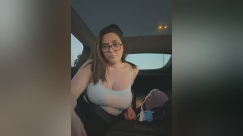 Video of a plus-sized woman with glasses, wearing a white tank top and black skirt, leaning in the backseat of a car, dimly lit interior.
