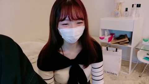 Video of an Asian woman with long dark hair and a white surgical mask, wearing a black-and-white striped top and a black scarf, sitting on a beige carpet in a minimalist room with a white dresser and shopping bags.