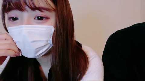 Video of a young Asian woman with straight, long brown hair, wearing a white face mask, pink eyeshadow, and a black jacket. She appears to be sneezing.