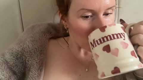 Video of a fair-skinned, brunette woman in a fluffy robe, sipping from a white ceramic mug adorned with red heart patterns. \"Mummy\" is written on the mug. Background is neutral and soft-focused.