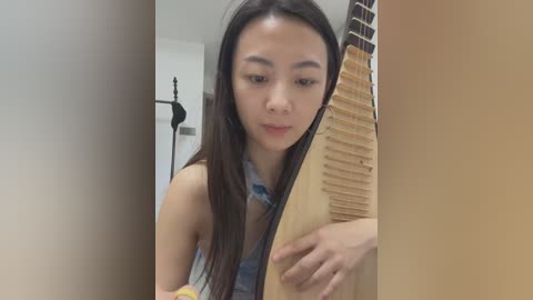 Video of an Asian woman with long black hair, wearing a denim shirt, playing a wooden guzheng in a modern room with beige walls.