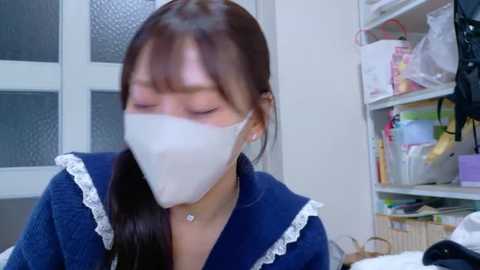 A video of an Asian woman with long brown hair, wearing a blue dress with white lace trim, and a white mask, seated indoors with a cluttered background.