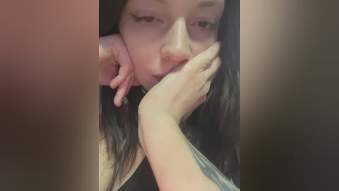 Media: Video of a woman with long black hair, pale skin, and prominent cheekbones, lying on her side, eyes half-closed, resting her chin on her hand, wearing a black top, in a dimly lit room.