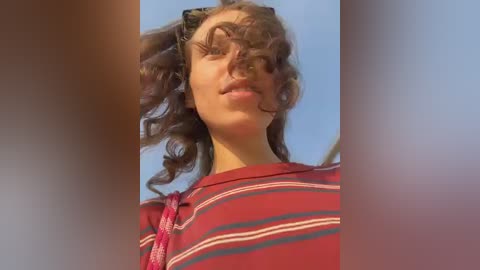 Video of a young woman with curly brown hair, wearing a red and white striped t-shirt, captured from a low angle, with a blurred background.
