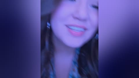 Blurry close-up video of a smiling young woman with light skin, wearing a floral-patterned top, set against a purple-toned background.