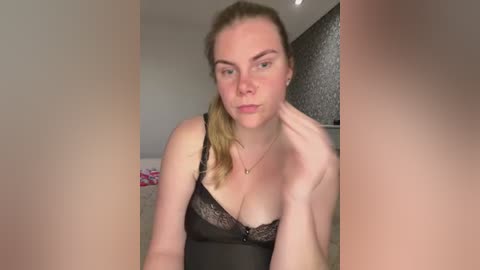 Video of a fair-skinned woman with blonde hair tied back, wearing a black lace bra, standing in a bedroom with grey patterned wallpaper and pink bedding visible.