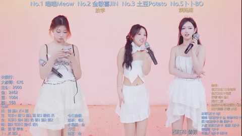 Video of three Asian women, all with fair skin, performing on stage. They wear white dresses and hold microphones, singing into them. The background is a gradient of pink and peach.