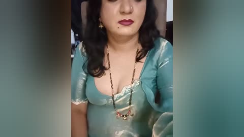 A video of a South Asian woman with fair skin, wearing a teal satin sari with a plunging neckline, gold earrings, and a necklace, against a blurred background.