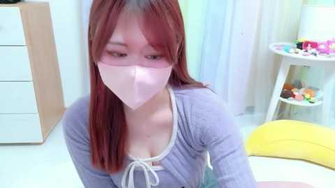 Video of a young Asian woman with straight red hair, wearing a pink surgical mask, light purple top, and white pants, sitting in a brightly lit room with pastel walls, toys, and a white dresser.