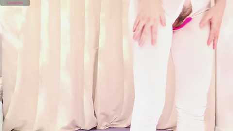 Video of a woman in white leggings, holding a pink vibrator against her crotch. She has fair skin and is standing in front of beige curtains.